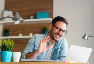 A white male wearing glasses, a white shirt and casual button up shirt smiling and waving to his computer screen while working in a home office.