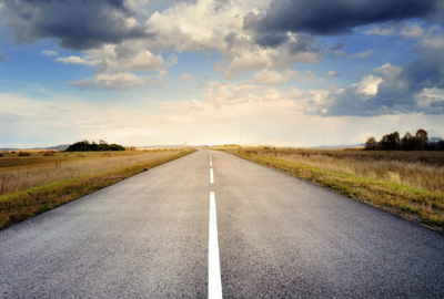 A view down the middle of a country road with the sun peeking out from behind the clouds.